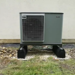 Zewnętrzna jednostka pompy ciepła Nibe F2040, umieszczona na czarnych podkładkach na żwirowym podłożu, z fragmentem trawnika i elewacji budynku w tle.