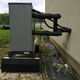 TERM-HOUSE WOJCIECH NOWAK - Szara pompa ciepła zamontowana na zewnątrz budynku z widocznymi rurami w izolacji, na tle zieleni i motocykla.