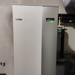 TERM-HOUSE WOJCIECH NOWAK - Biała pompa ciepła NIBE z podłączonymi rurami w kotłowni, widoczne elementy instalacji hydraulicznej i zbiornik wyrównawczy.