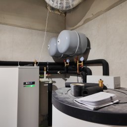 TERM-HOUSE WOJCIECH NOWAK - Biała pompa ciepła NIBE z zielonym wskaźnikiem pracy, zbiornik akumulacyjny, dwa szare naczynia wzbiorcze, izolowane rury, manometry, oraz dokumentacja na zbiorniku w surowym pomieszczeniu.