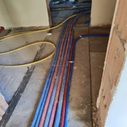 Wiązka niebiesko-czerwonych rur instalacyjnych ułożona na szarej posadzce w surowym stanie, widoczne fragmenty ścian w trakcie wykończenia i żółta rura w tle.