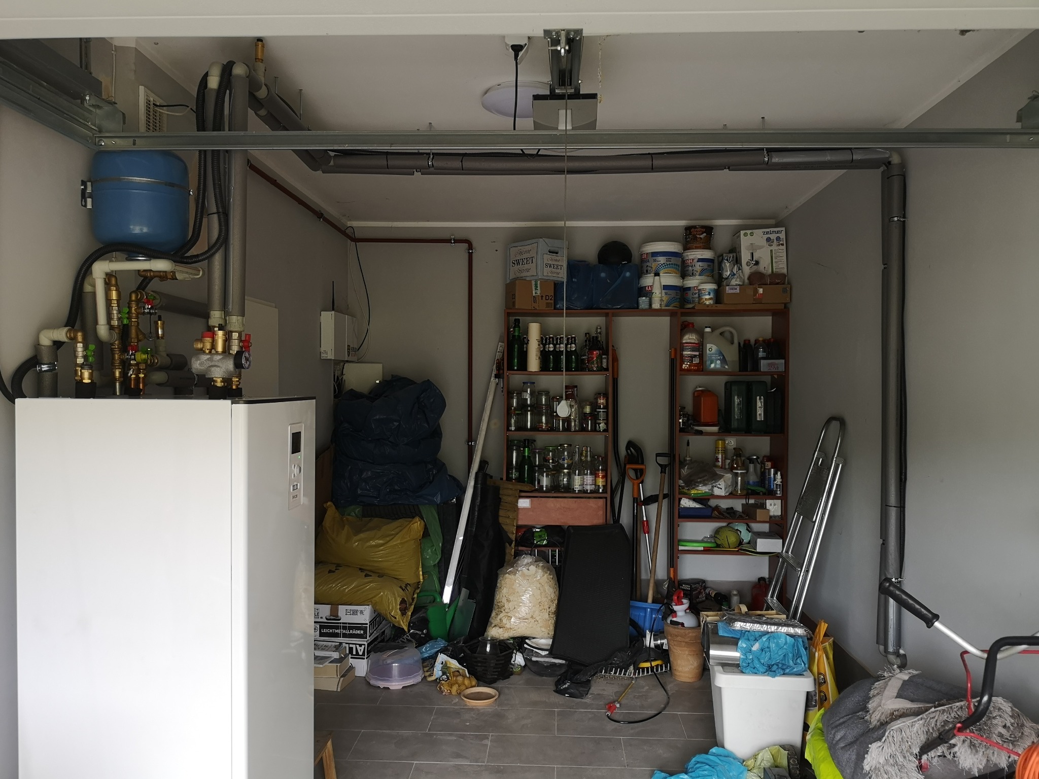 Biała pompa ciepła z niebieskim zbiornikiem wyrównawczym i orurowaniem w garażu z regałami wypełnionymi narzędziami i różnymi przedmiotami.
