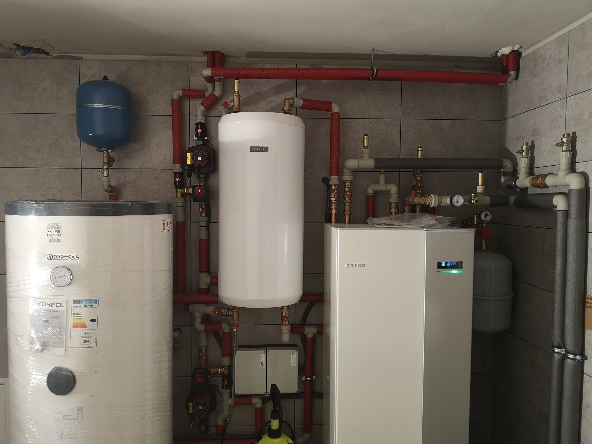 Kotłownia z zbiornikami Kospel i Nibe, instalacją hydrauliczną z czerwonymi rurami i szarą glazurą na ścianach.
