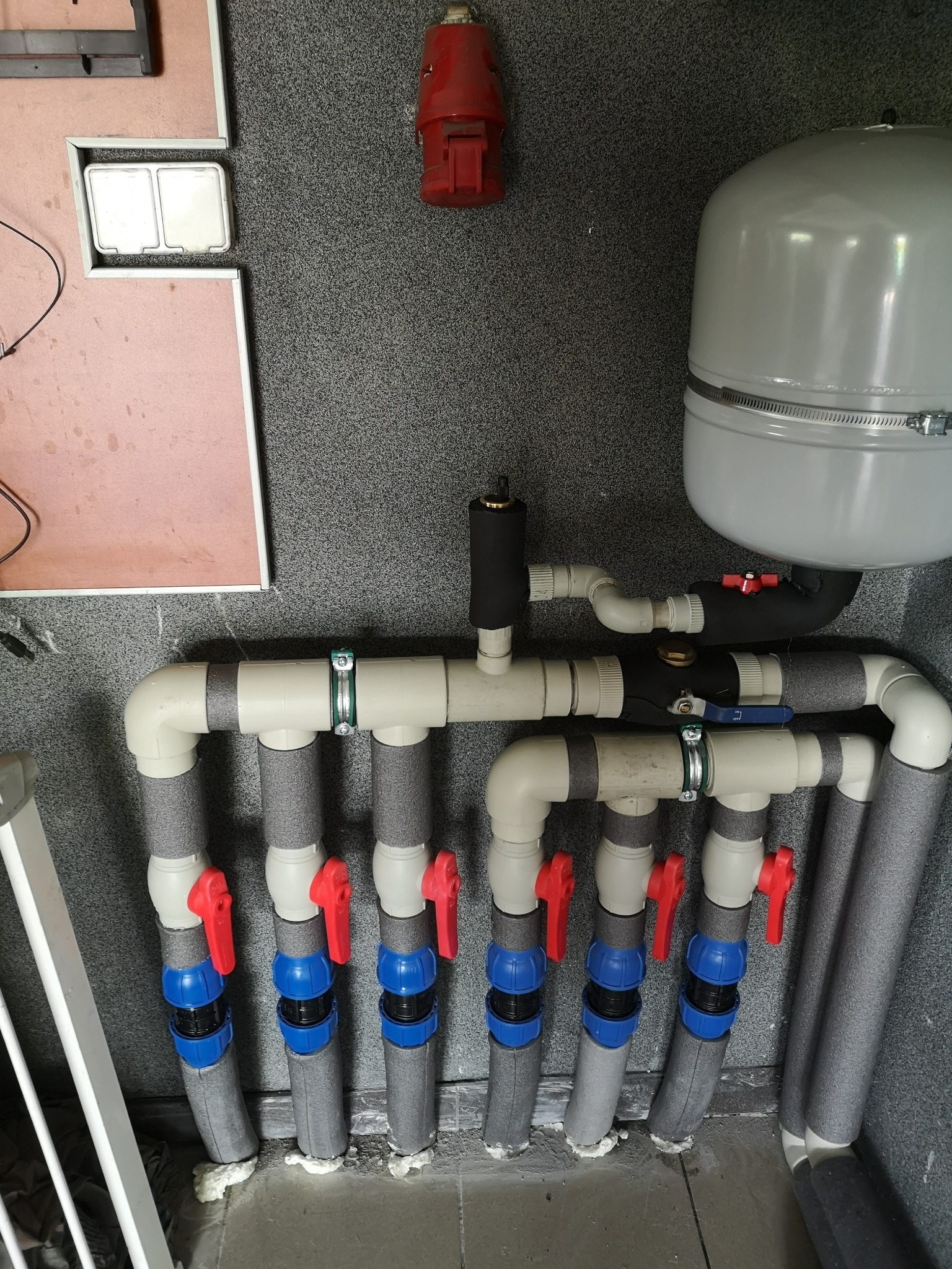 Szereg rur instalacji grzewczej z zaworami odcinającymi, izolacją termiczną i zbiornikiem wyrównawczym na tle szarej ściany i fragmentu białej balustrady.