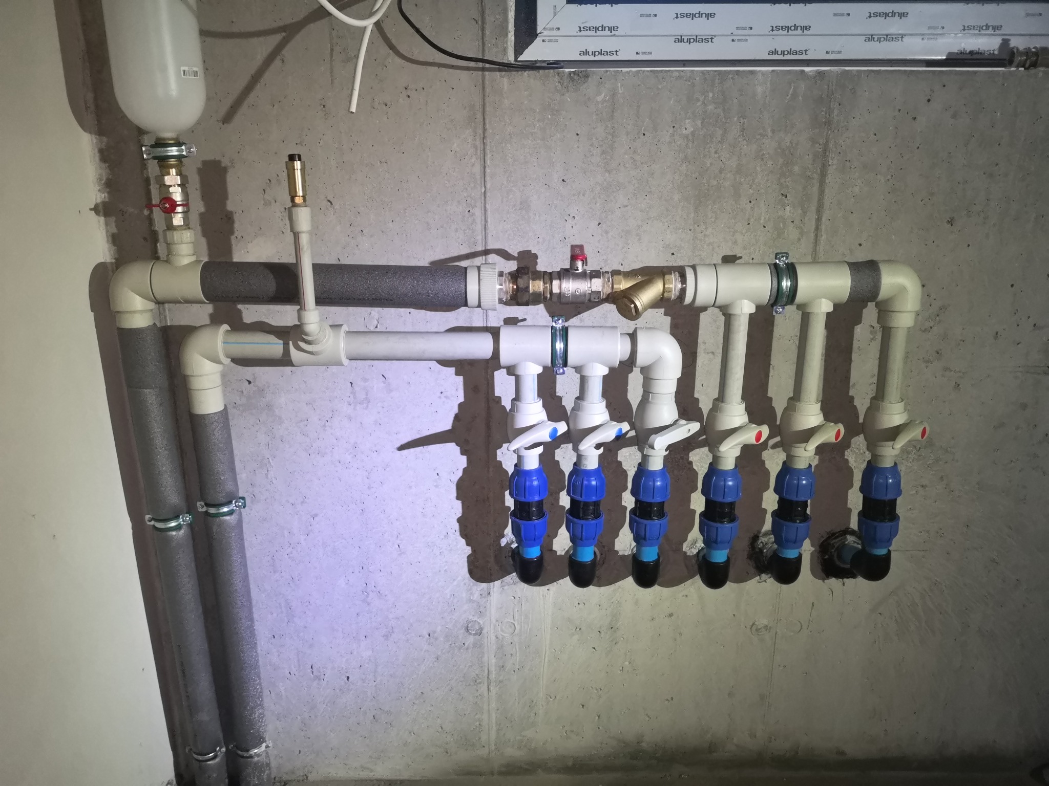 Rozdzielacz instalacji wodnej z sześcioma zaworami odcinającymi, zamontowany na surowej ścianie z betonu komórkowego, z widocznymi rurami w izolacji termicznej.