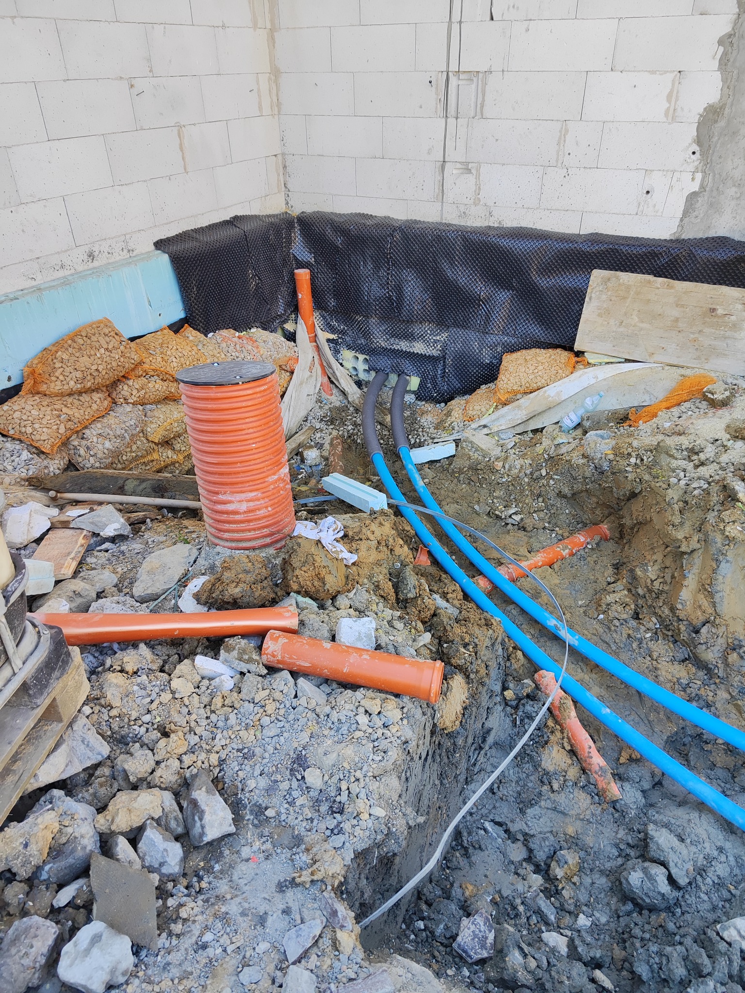 Wykop z widocznymi niebieskimi rurami instalacji wodnej, pomarańczowymi rurami kanalizacyjnymi, zasypywanymi workami z kruszywem i zabezpieczeniem fundamentów budynku w trakcie budowy.
