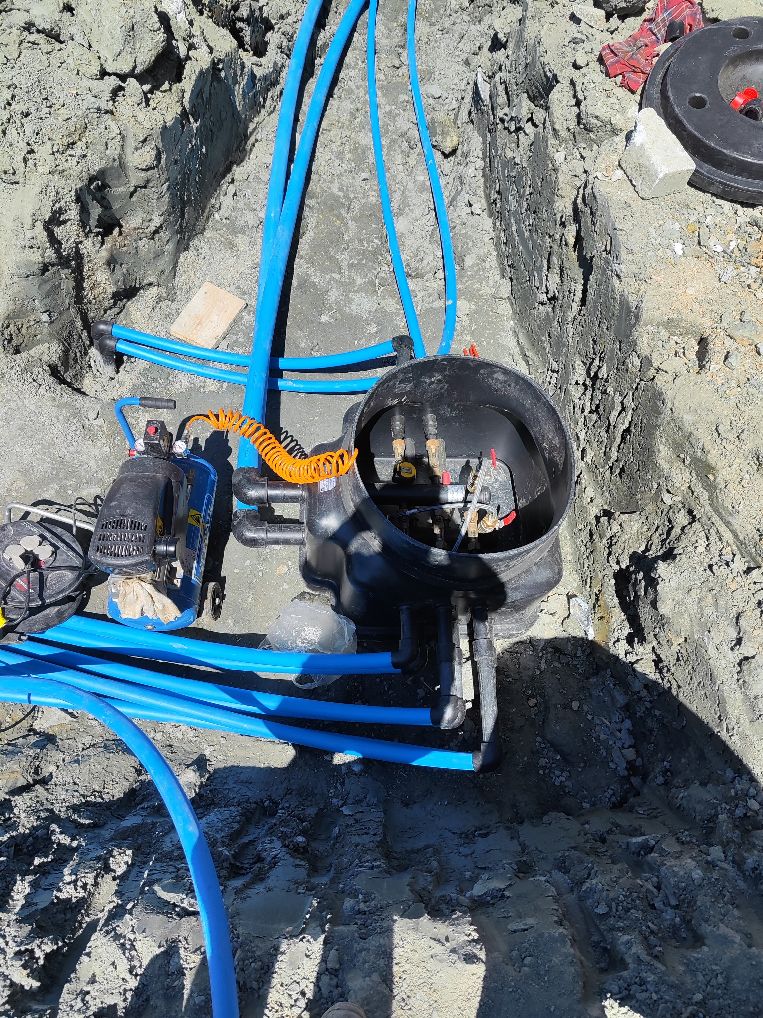 Instalacja hydrauliczna w wykopie ziemnym z widocznym kompresorem, niebieskimi rurami i czarną skrzynką z zaworami.