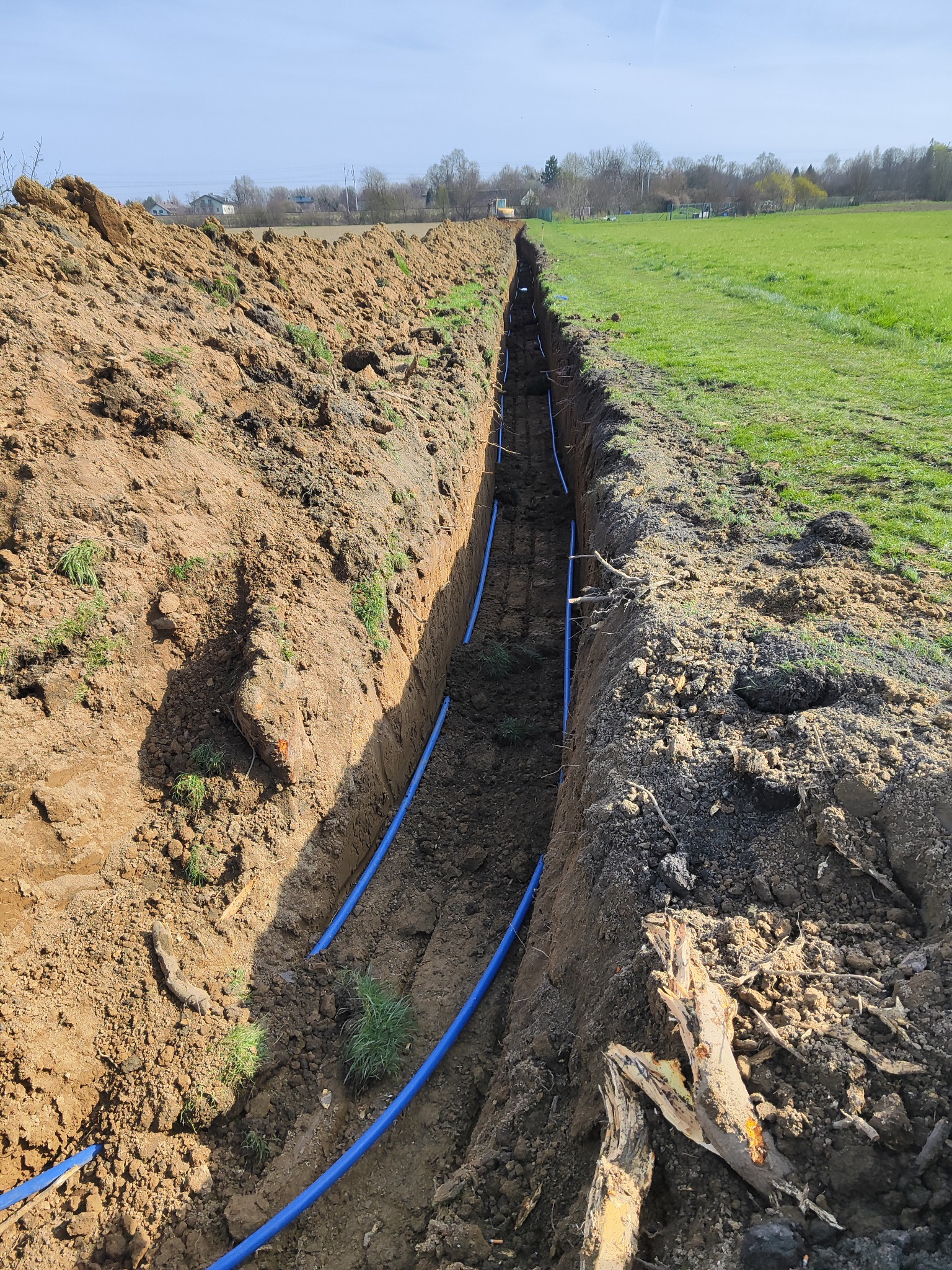 Wykop w ziemi z ułożonymi niebieskimi rurami, widoczny krajobraz z drzewami i polem w tle, ziemia na brzegach wykopu z kępami trawy.