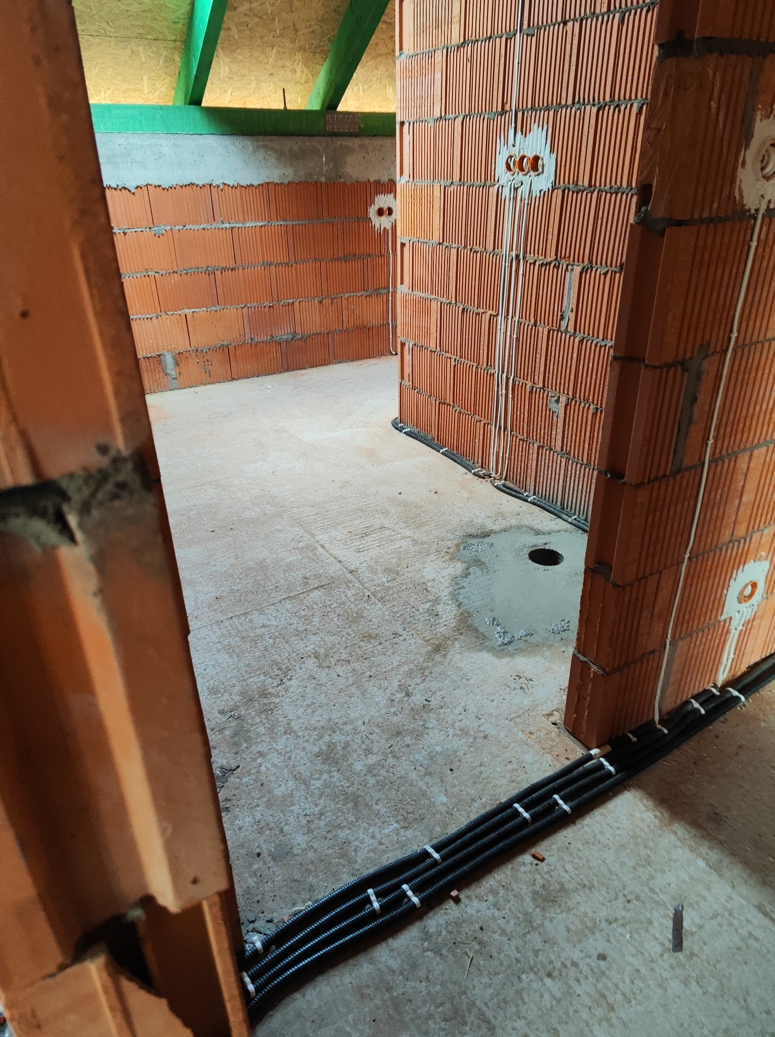 Wnętrze w trakcie budowy z ceglanymi ścianami z widocznymi przewodami elektrycznymi i rurami instalacji grzewczej układanymi wzdłuż podłogi.