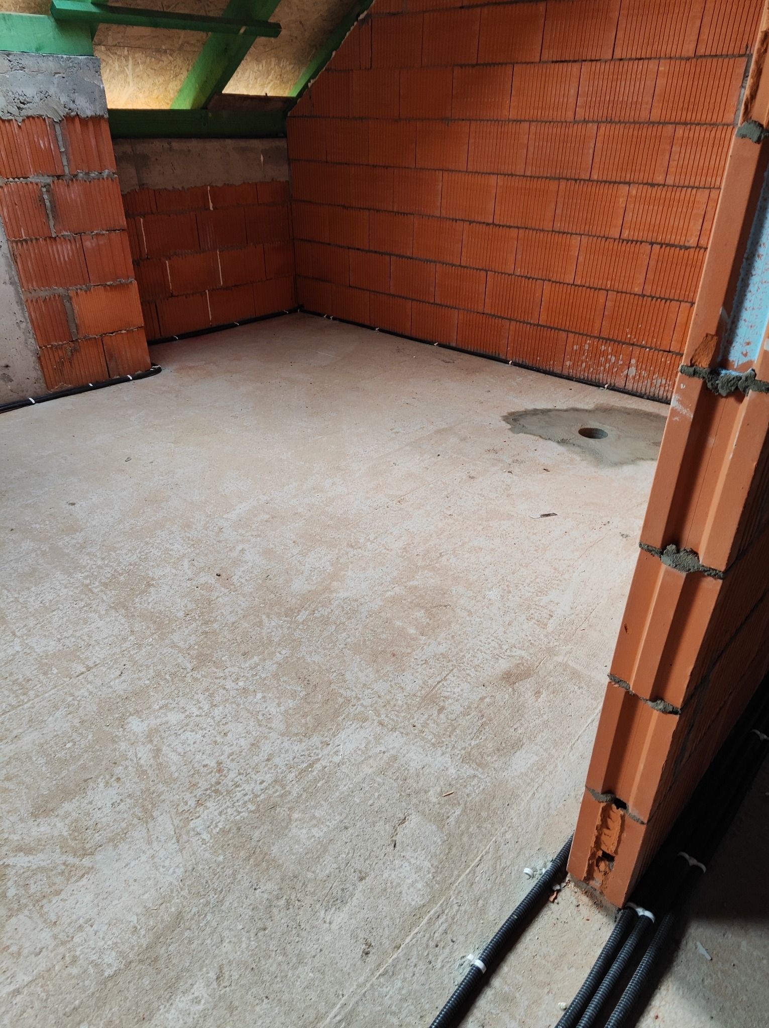 Surowy stan poddasza z częściowo wymurowanymi ścianami z czerwonej cegły, widoczne czarne rury instalacyjne przy podłodze i otwór w wylewce, przygotowane do dalszych prac wykończeniowych.