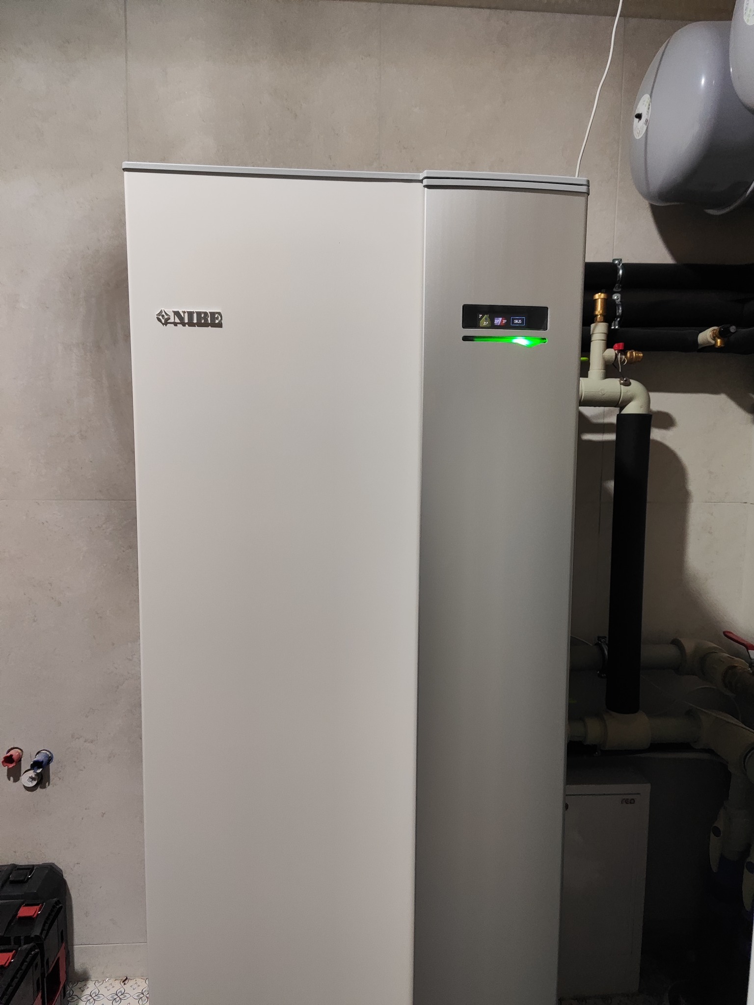 Biała pompa ciepła NIBE z podłączonymi rurami w kotłowni, widoczne elementy instalacji hydraulicznej i zbiornik wyrównawczy.