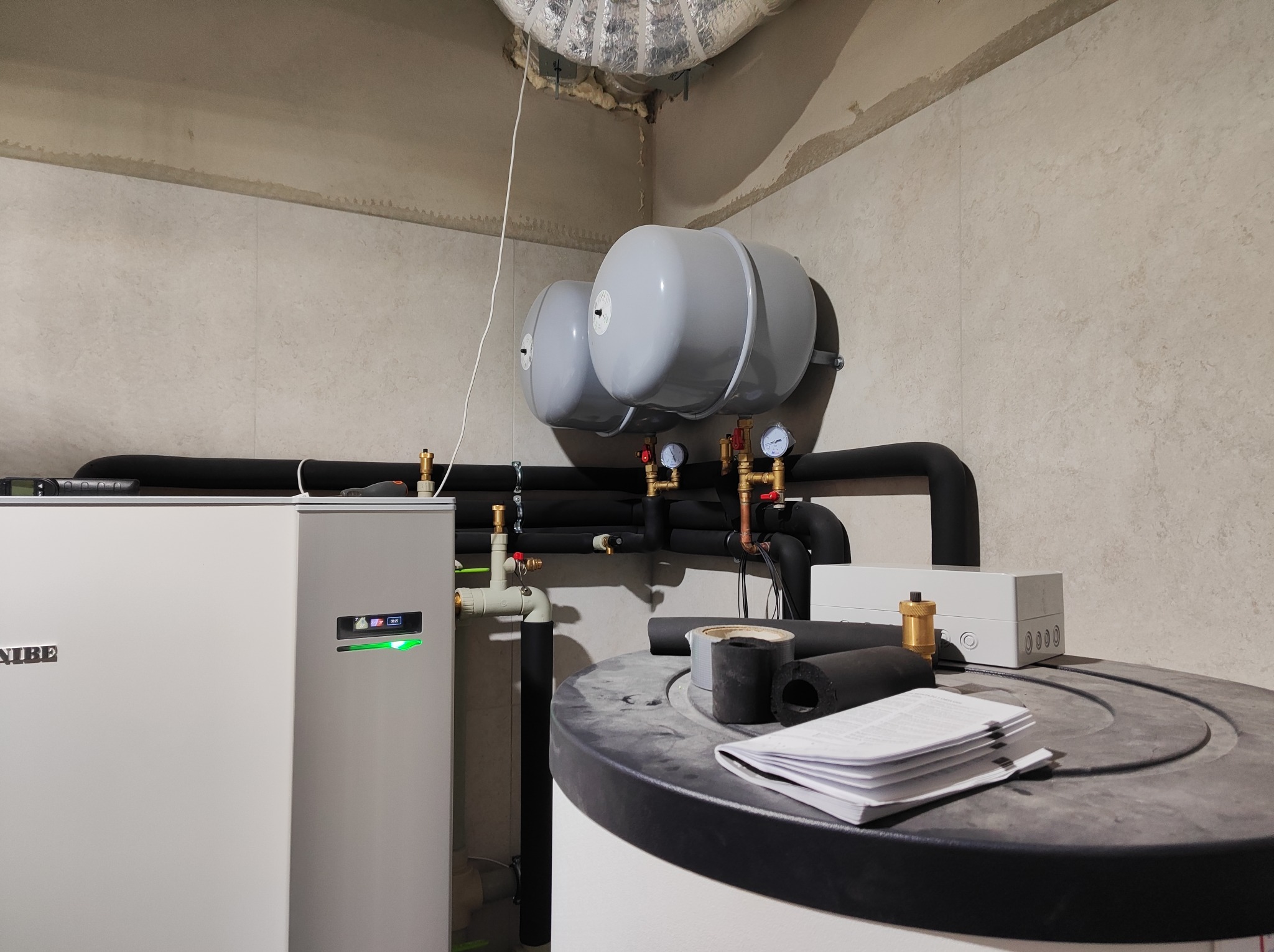 Biała pompa ciepła NIBE z zielonym wskaźnikiem pracy, zbiornik akumulacyjny, dwa szare naczynia wzbiorcze, izolowane rury, manometry, oraz dokumentacja na zbiorniku w surowym pomieszczeniu.