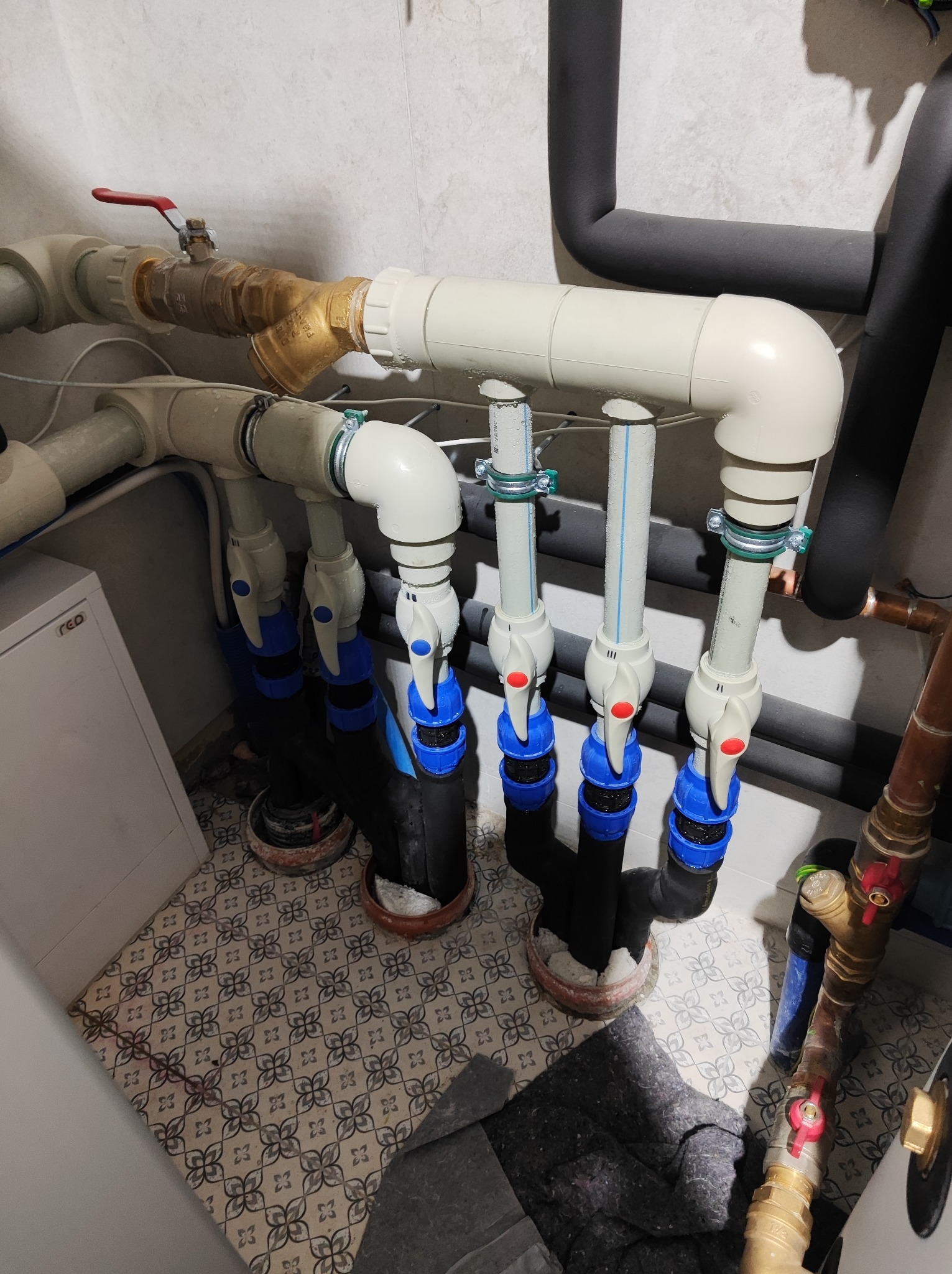 Złożony układ rur z zaworami i izolacją termiczną, widoczne liczne połączenia i elementy instalacji w pomieszczeniu z wzorzystą podłogą.