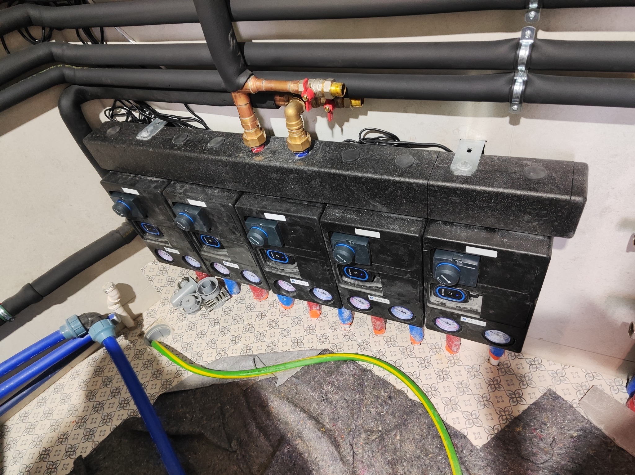 Rozdzielacz ogrzewania podłogowego z manometrami, rurami PEX i przewodami elektrycznymi, widok z góry na tle wzorzystej podłogi i zaizolowanych rur grzewczych.