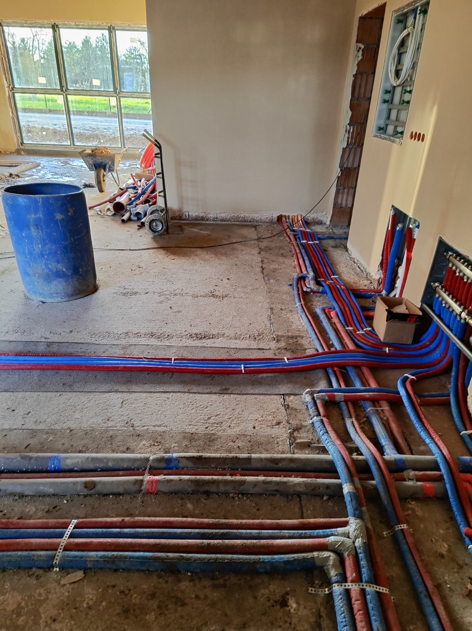 Instalacja hydrauliczna w surowym stanie: liczne czerwono-niebieskie rury prowadzone po podłodze, z widocznym rozdzielaczem i elementami montażowymi, w tle widoczne okno i narzędzia budowlane.