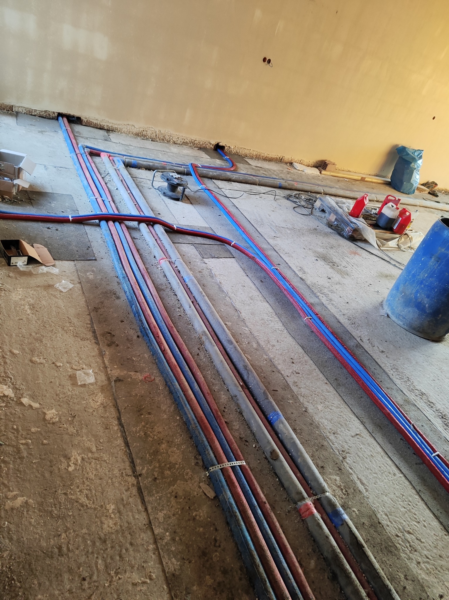 Instalacja rur grzewczych i wodnych w bruzdach w podłodze podczas remontu, widoczne narzędzia i materiały budowlane.