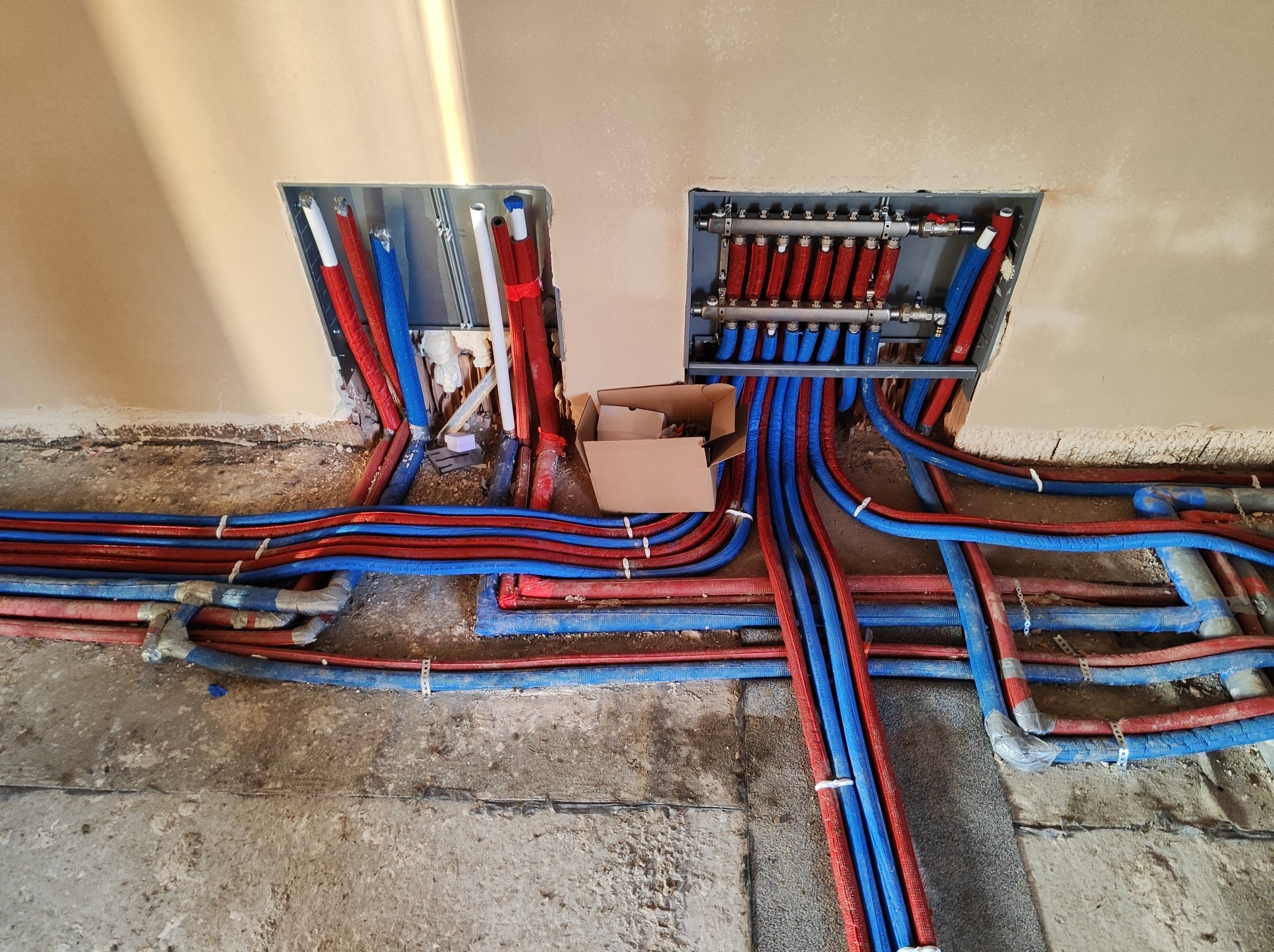 Rozdzielacz ogrzewania podłogowego w trakcie montażu w Głębowicach, widoczne rury doprowadzające ciepłą i zimną wodę, surowy stan posadzki przed wylewką.