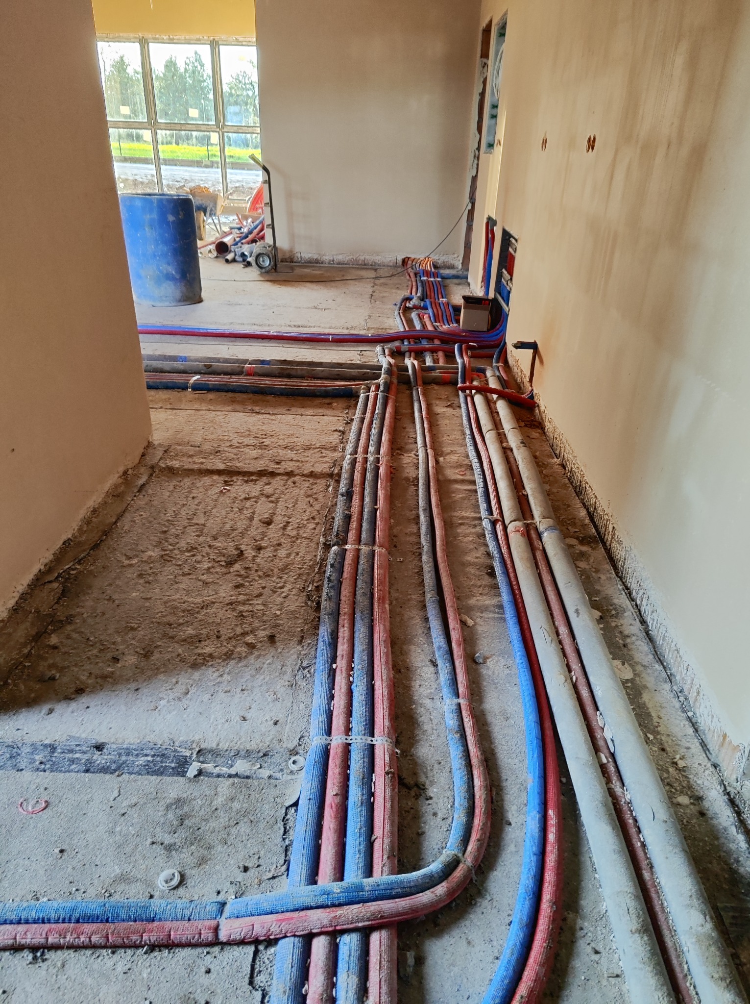 Wnętrze w trakcie remontu z widocznymi rzędami rur instalacyjnych w kolorach niebieskim, czerwonym i szarym ułożonymi na podłodze, z nieotynkowanymi ścianami i oknem w tle.
