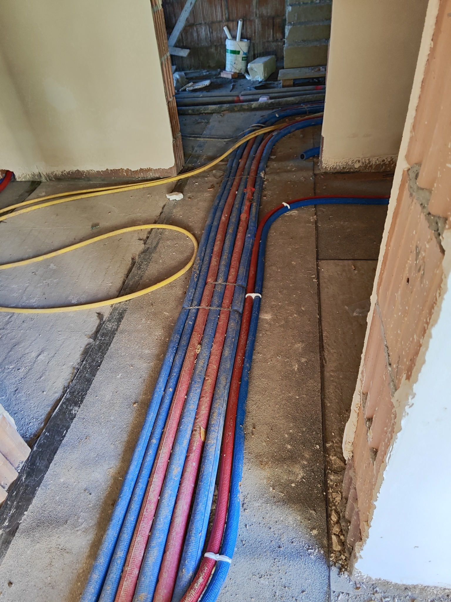 Wiązka niebiesko-czerwonych rur instalacyjnych ułożona na szarej posadzce w surowym stanie, widoczne fragmenty ścian w trakcie wykończenia i żółta rura w tle.