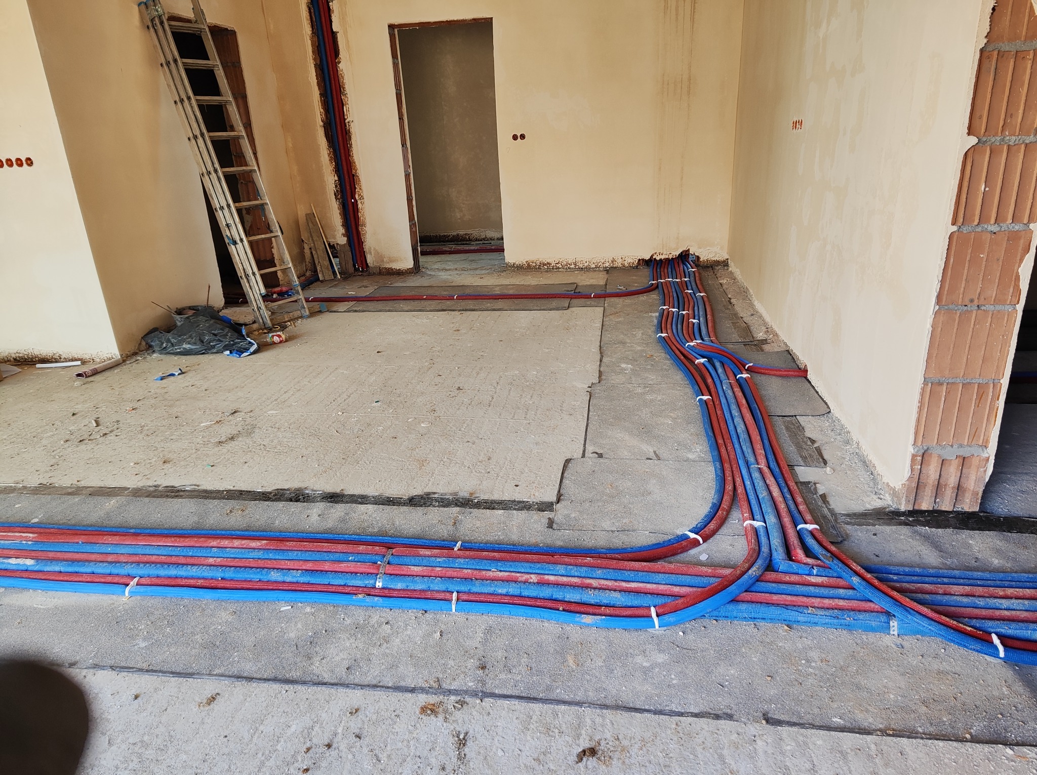 Instalacja rur grzewczych i wodnych w surowym stanie, widoczne rury w kolorze czerwonym i niebieskim ułożone na podłodze, w tle drabina oparta o ścianę i surowe ściany z widocznymi otworami...
