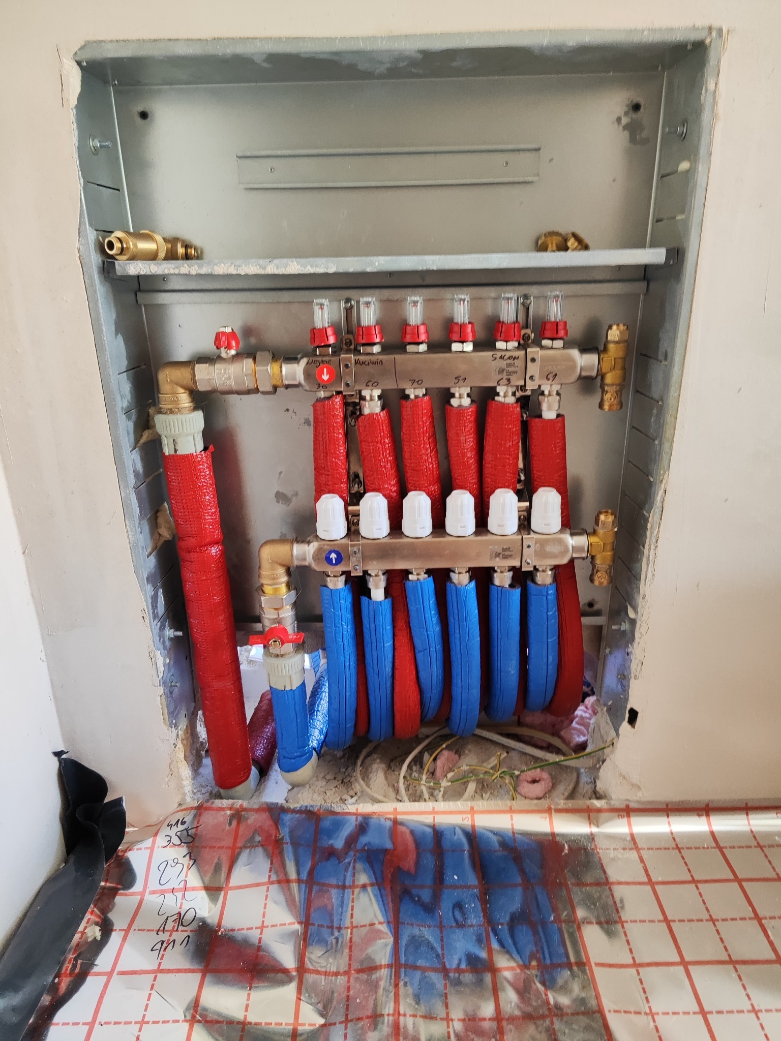 Rozdzielacz ogrzewania podłogowego w metalowej obudowie, z rurami w izolacji czerwonej i niebieskiej, widoczne zawory i manometry, na podłodze folia z nadrukowaną siatką i odręcznymi notatkami.