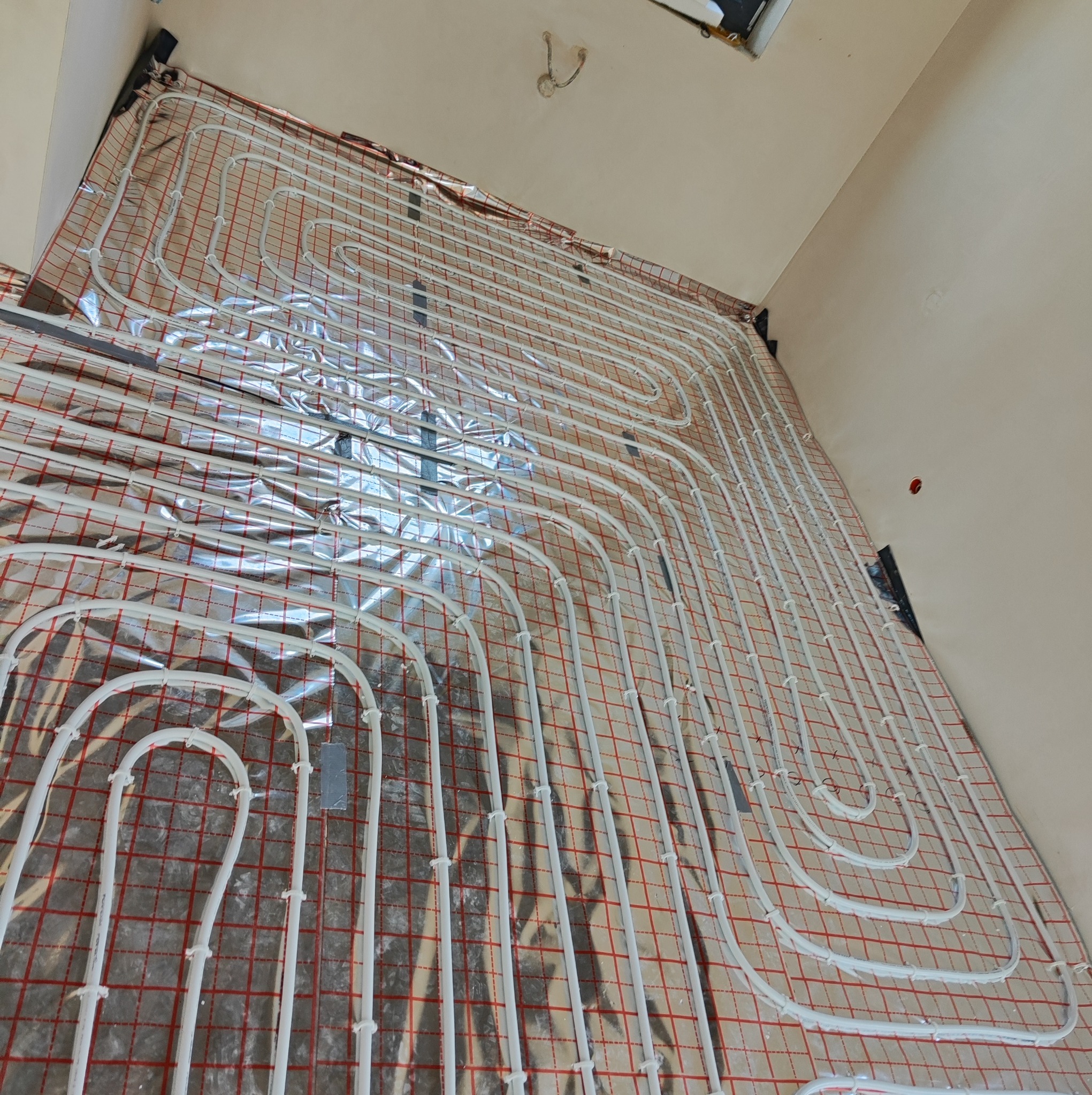 Białe rury ogrzewania podłogowego ułożone w pętle na izolacji z siatką w czerwono-białą kratkę, widok z góry pod kątem.