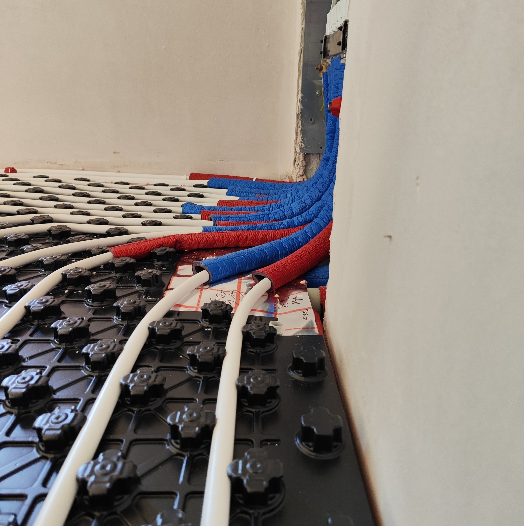Montaż ogrzewania podłogowego: białe rurki ułożone na czarnej macie z uchwytami, czerwono-niebieskie izolowane rury wchodzące w ścianę, widok z dołu.