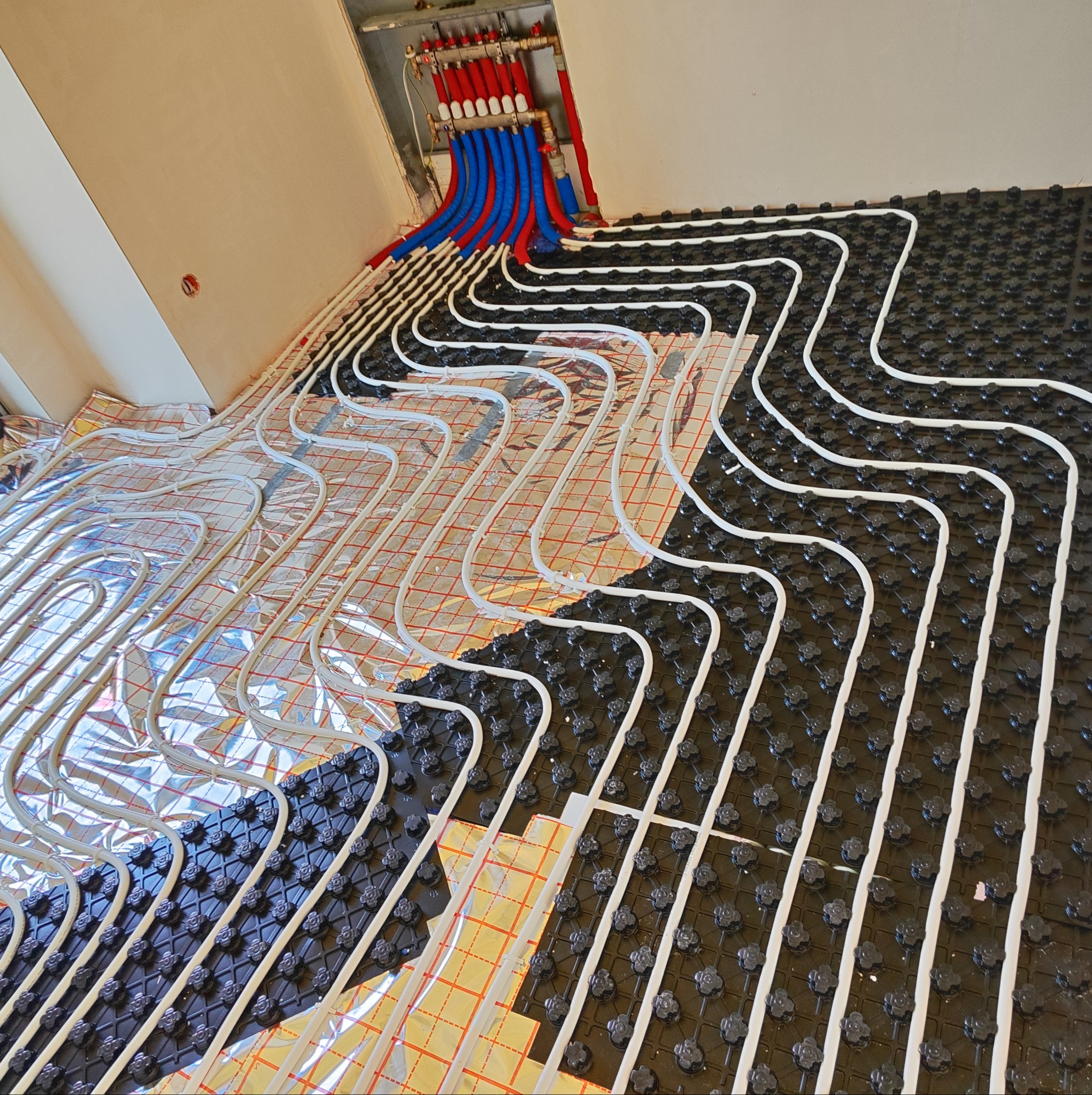 Rozłożona instalacja ogrzewania podłogowego z rurkami na folii aluminiowej i czarnej macie z uchwytami, widoczny rozdzielacz z czerwonymi i niebieskimi przewodami.
