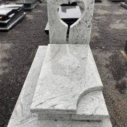 Jasnoszary, polerowany pomnik z granitu z wyciętym sercem, ustawiony na żwirowym podłożu wśród innych nagrobków.