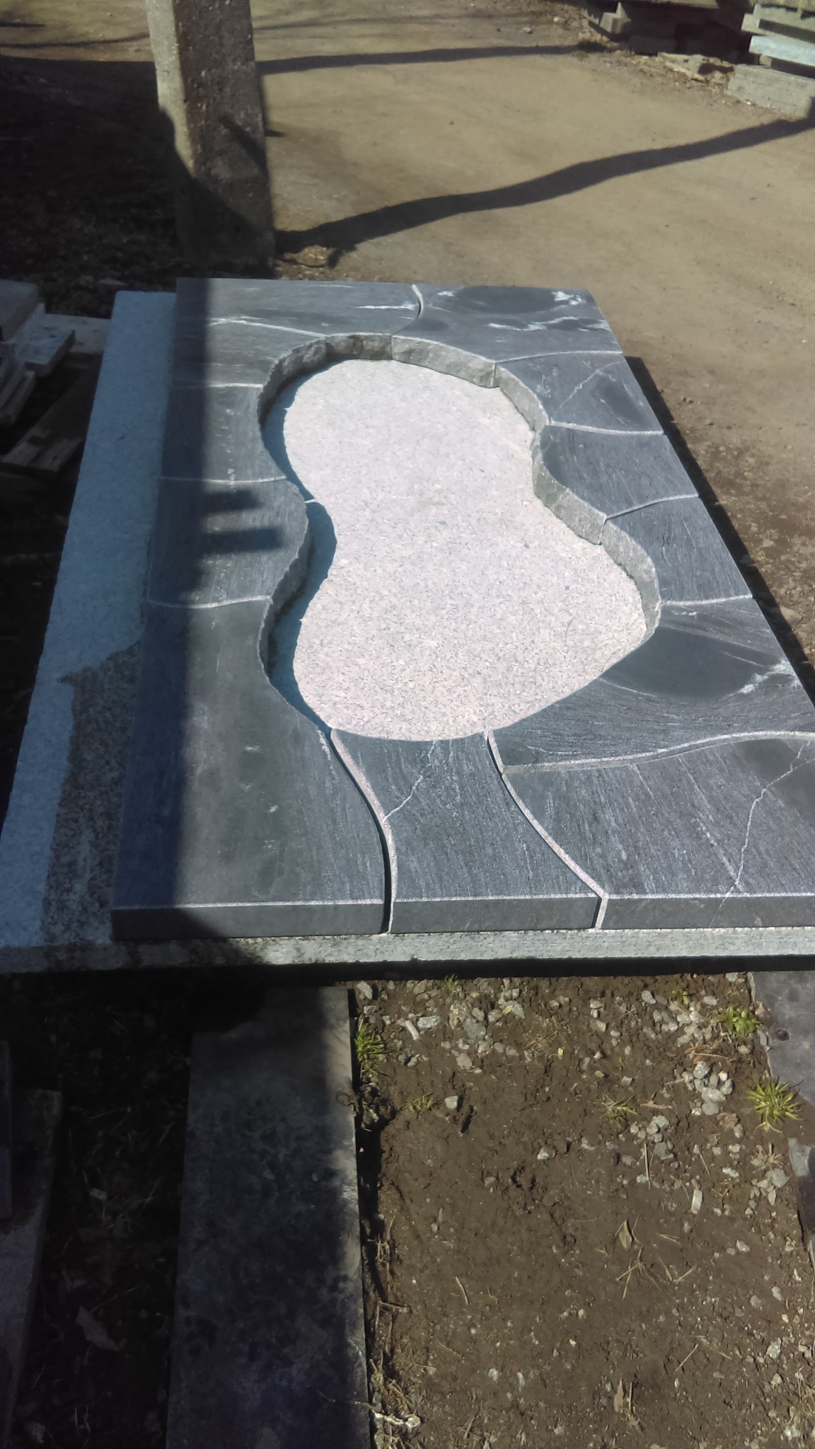 Surowy, częściowo obrobiony element nagrobka z ciemnoszarego kamienia z jasnoszarym wypełnieniem, leżący na ziemi w warsztacie kamieniarskim.