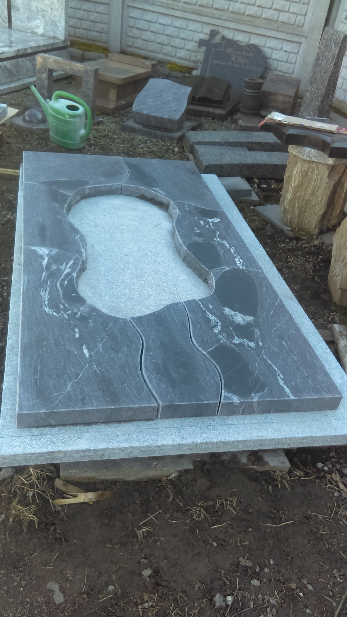 Element nagrobka z ciemnego kamienia z jasnymi żyłkami, z wyciętym otworem, leżący na jasnym kamiennym podłożu, na tle innych elementów nagrobnych i zielonej konewki.