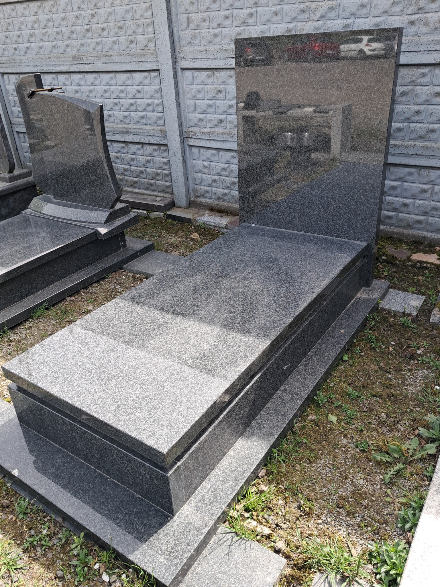 Nowoczesny, ciemnoszary pomnik z granitu z prostokątną płytą nagrobną i pionową tablicą, odbicie otoczenia na polerowanej powierzchni, ustawiony na terenie z ziemią i zielenią, obok betonowego...