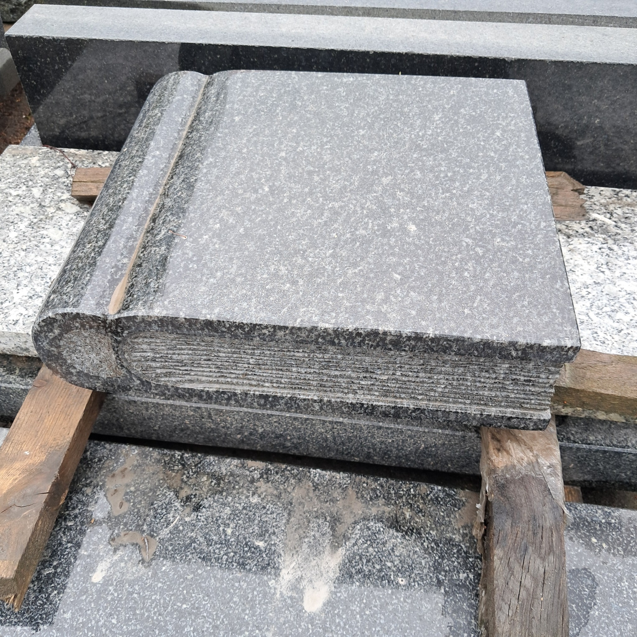 Szary, granitowy nagrobek w kształcie otwartej księgi, oparty na drewnianych legarach, z widoczną strukturą kamienia i imitacją kartek.