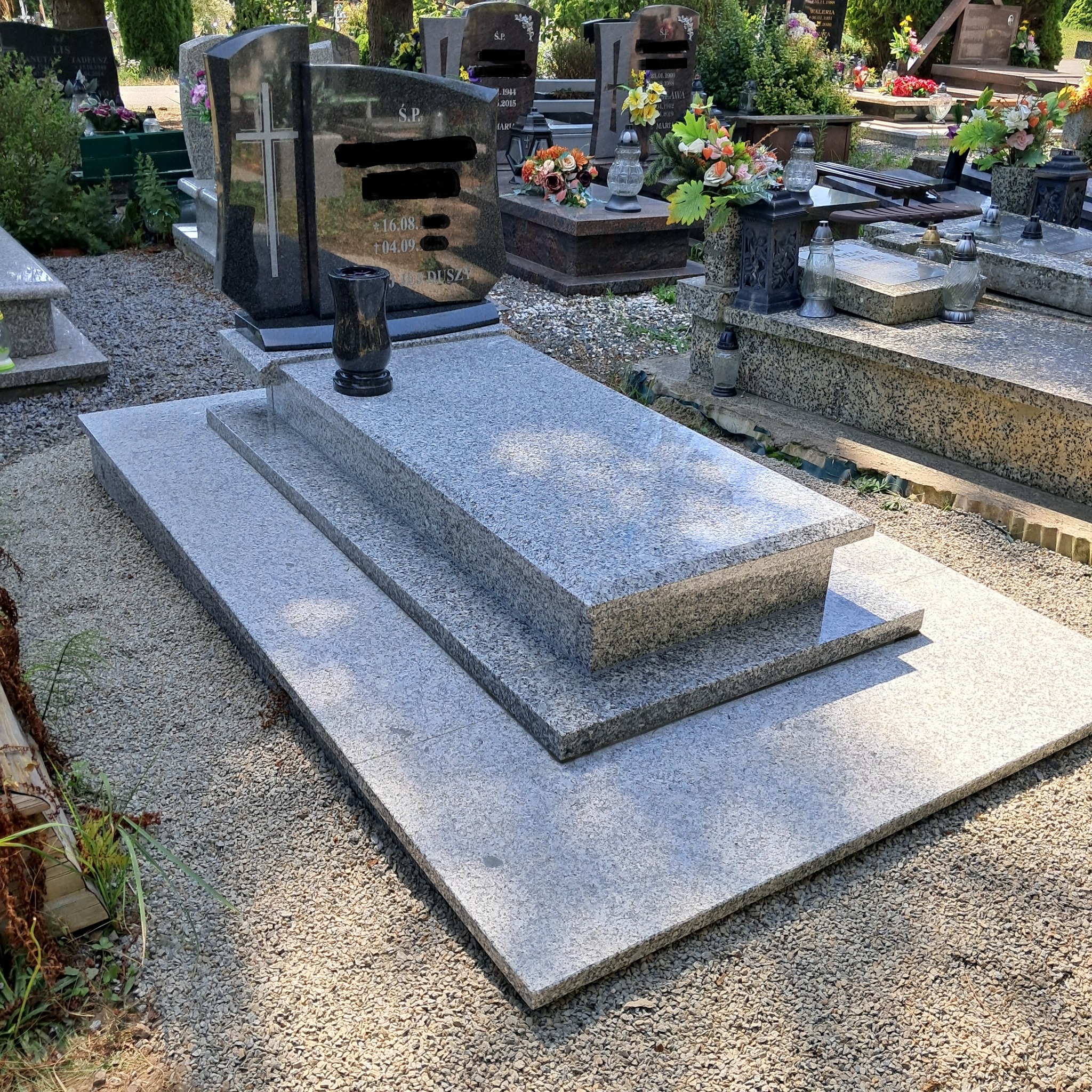 Granitowy nagrobek z trzema poziomami, otoczony żwirem na cmentarzu, w tle inne groby i zieleń.