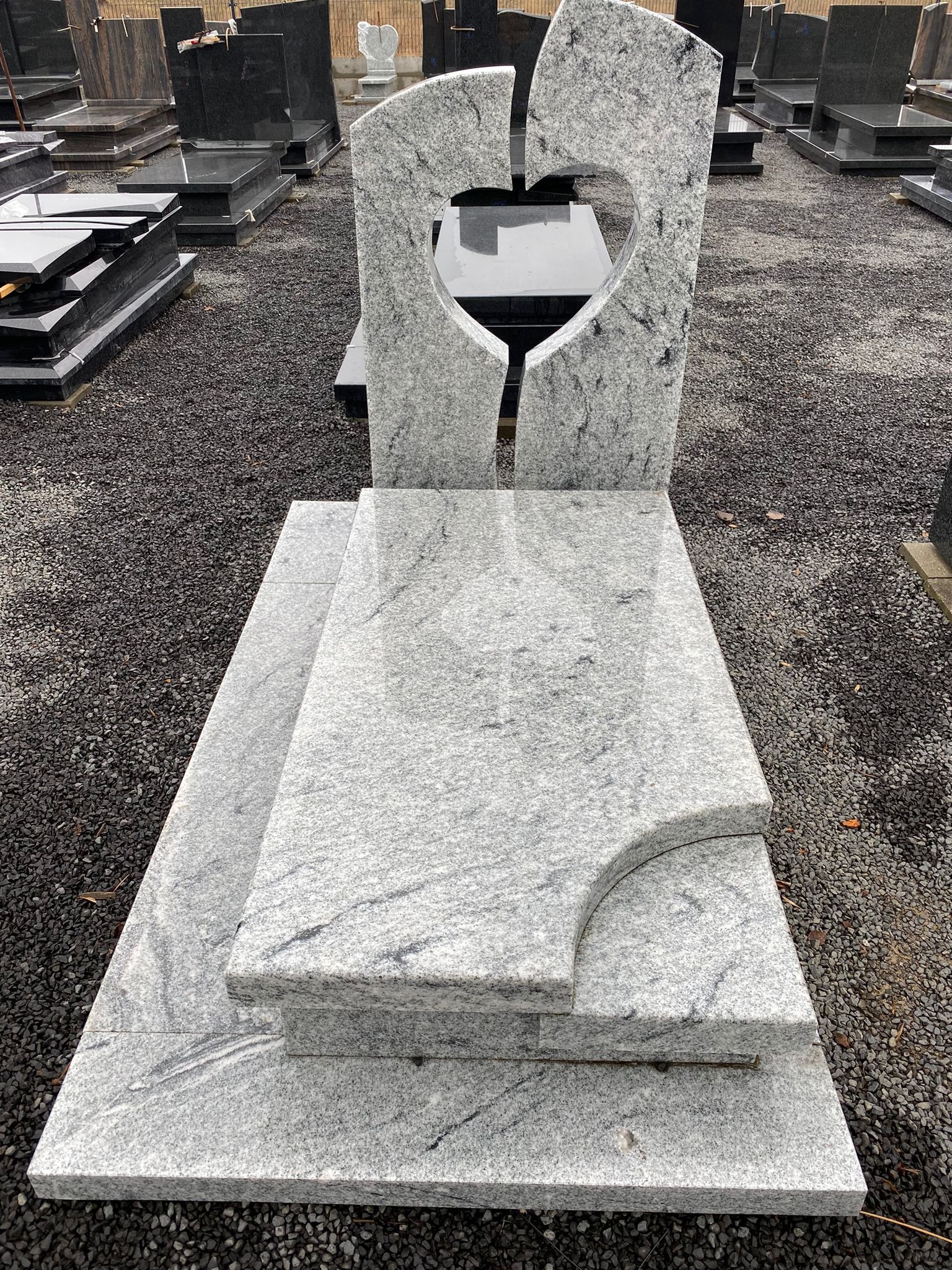 Jasnoszary, polerowany pomnik z granitu z wyciętym sercem, ustawiony na żwirowym podłożu wśród innych nagrobków.