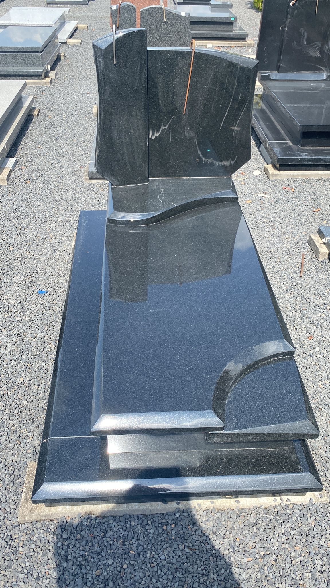 Czarny, błyszczący pomnik z granitu o nowoczesnym kształcie, z widocznymi zarysowaniami na powierzchni, ustawiony na żwirowym podłożu wśród innych nagrobków.