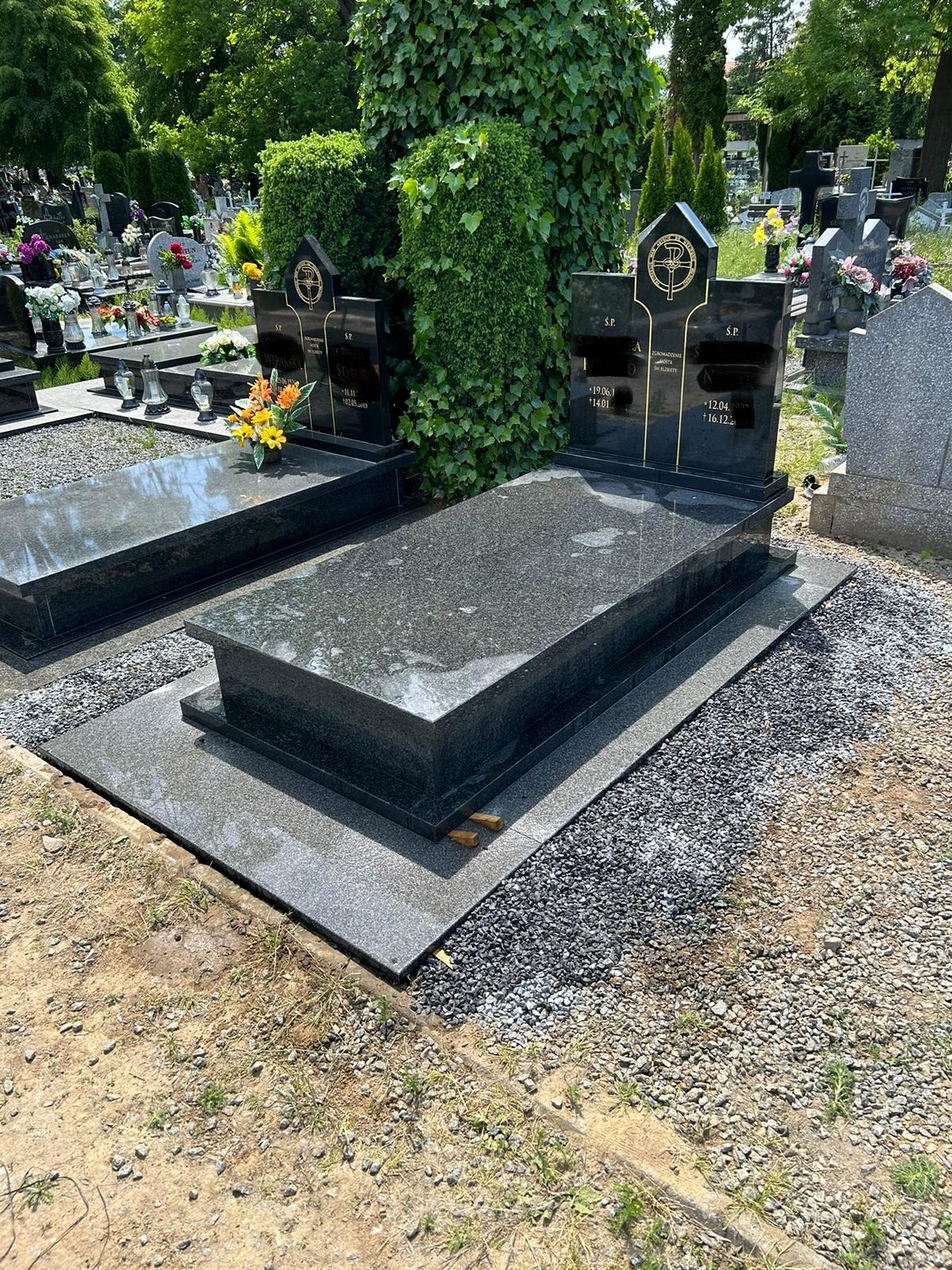Ciemny, granitowy pomnik nagrobny na cmentarzu, otoczony drobnym żwirem i zielenią. Widoczne kwiaty w wazonie. Dwa drewniane kliny pod płytą.