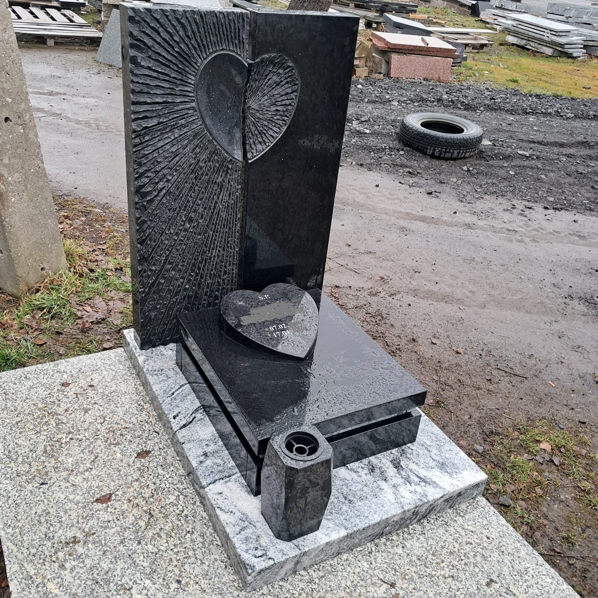 Czarny, granitowy pomnik z wyrytym sercem i małą tabliczką w kształcie serca z datami, ustawiony na szarej, kamiennej podstawie, w tle inne elementy kamienne i opona.