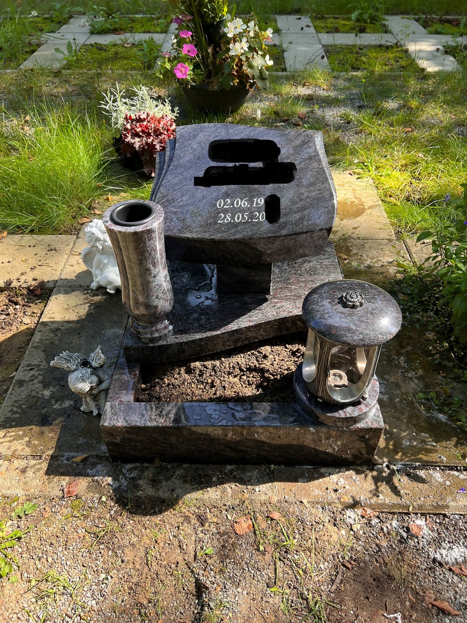 Wielopoziomowy pomnik z ciemnego granitu z wyrytymi datami, wazonem i lampionem, ozdobiony figurkami aniołów, otoczony zielenią i kwiatami na cmentarzu.