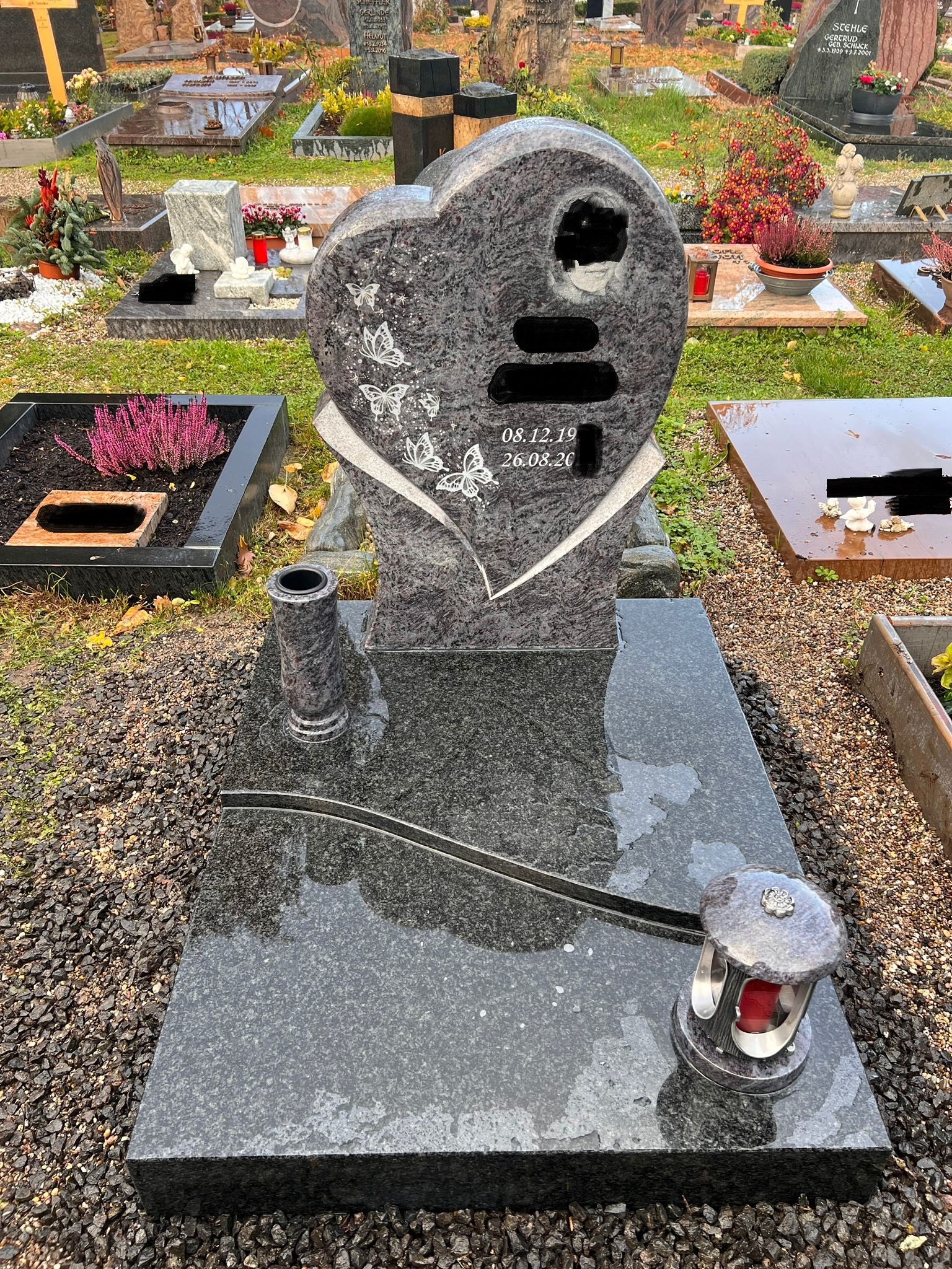 Granitowy nagrobek w kształcie serca z wygrawerowanymi motylami i datami, wazon i lampion na płycie, na cmentarzu.