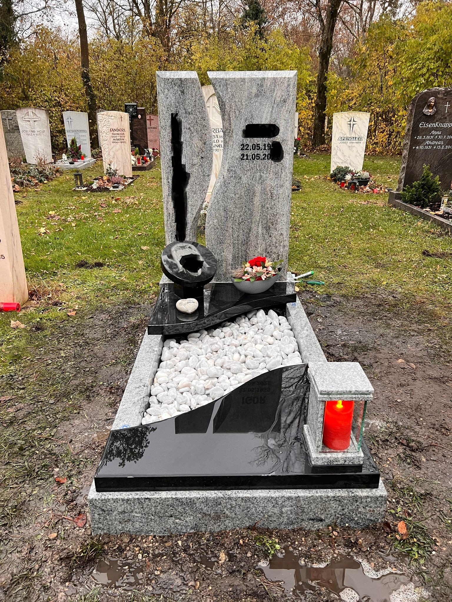 Nowoczesny nagrobek z szarego granitu z czarnymi elementami i białymi kamykami dekoracyjnymi, na cmentarzu z widocznymi innymi nagrobkami i drzewami w tle.