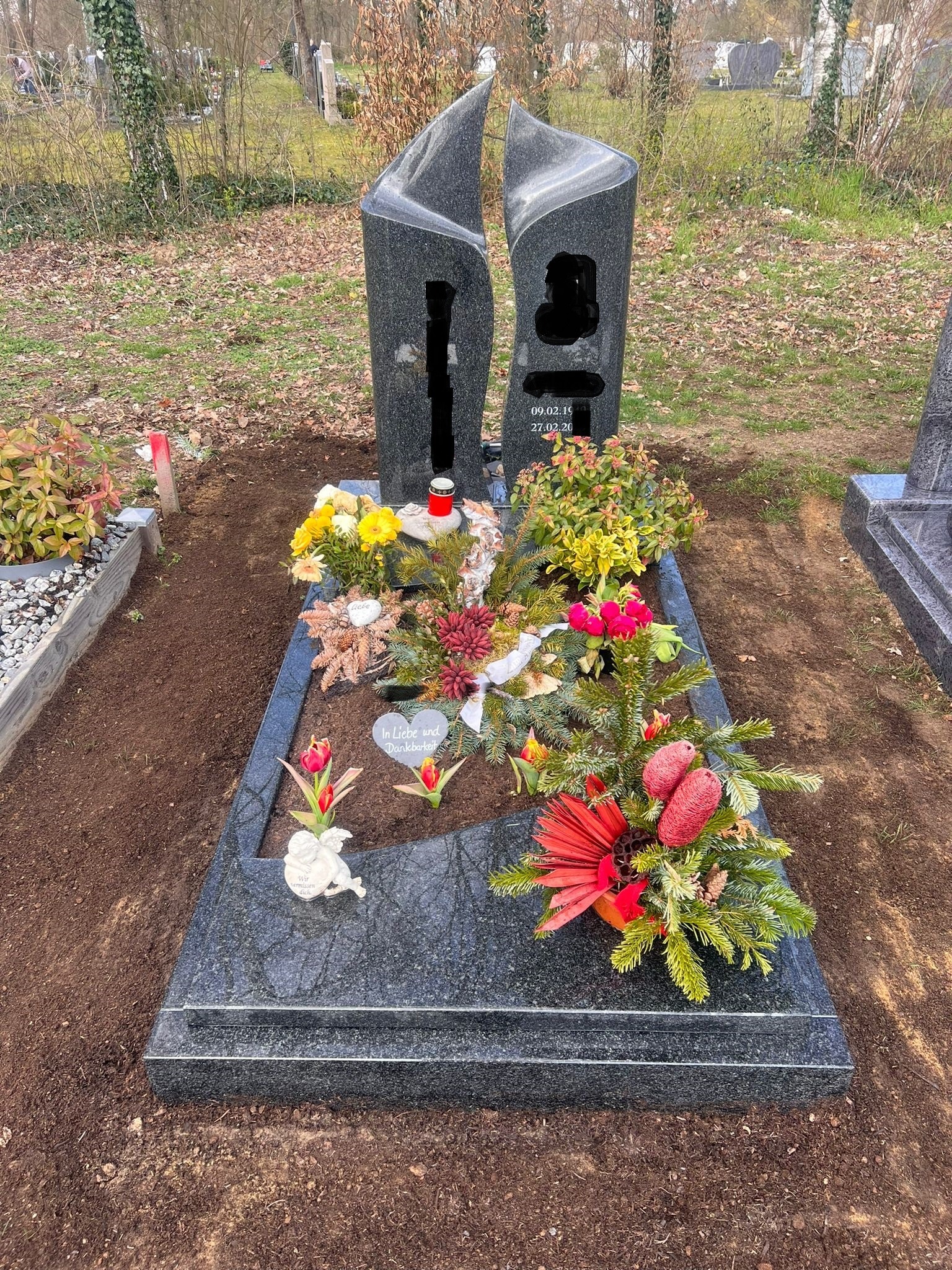 Granitowy pomnik z dwoma pionowymi elementami w kształcie płomieni, udekorowany kwiatami i dekoracjami świątecznymi na cmentarzu.