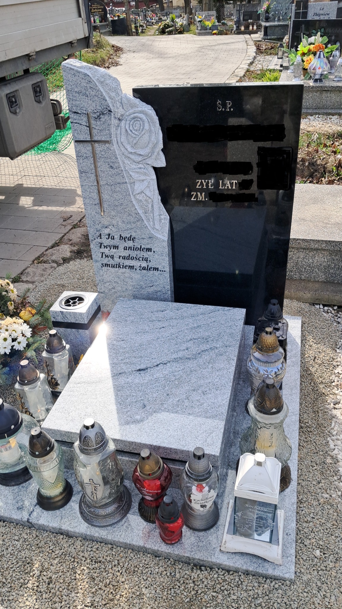Granitowy pomnik z rzeźbioną różą i krzyżem, otoczony zniczami na żwirowym podłożu cmentarza.