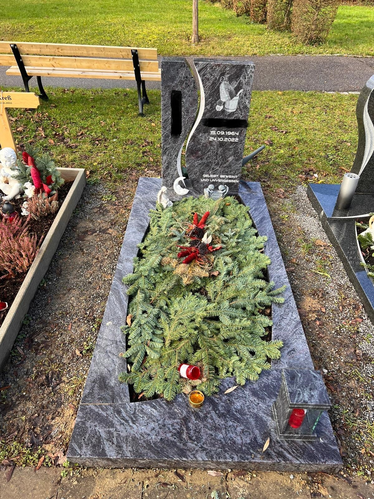 Granitowy pomnik z wygrawerowanym motywem i datami, udekorowany zielonymi gałązkami, czerwonymi ozdobami i lampionami, na cmentarzu.