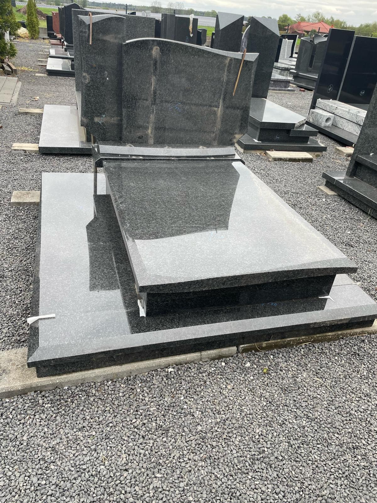 Czarny, polerowany nagrobek z granitu, składający się z płyty podstawowej, nakrywy i pionowej tablicy, prezentowany na placu z szarego żwiru.
