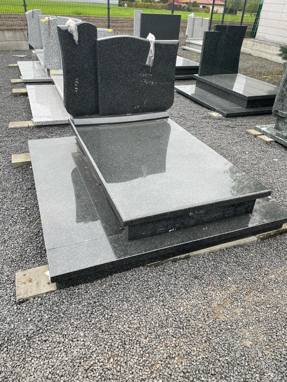 Nowy, czarny, granitowy pomnik nagrobny, ustawiony na betonowych blokach na żwirowym placu, z innymi nagrobkami w tle.