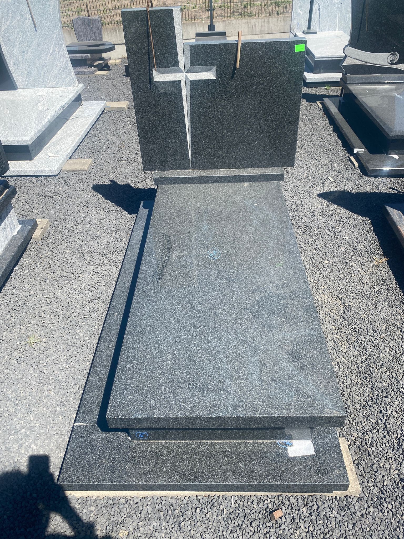 Granitowy nagrobek z krzyżem na tle szarego żwiru, widoczne inne nagrobki w tle