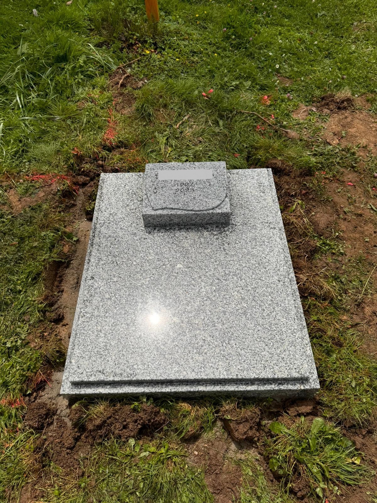 Jasnoszary, granitowy pomnik nagrobny z wygrawerowanym napisem, otoczony świeżą ziemią i trawą, w trakcie montażu.