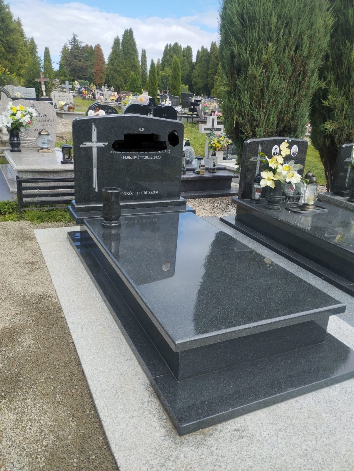 Nowoczesny, czarny, dwupoziomowy pomnik z ciemnego kamienia na cmentarzu, z wygrawerowanym krzyżem i inskrypcją, otoczony zielenią drzew.