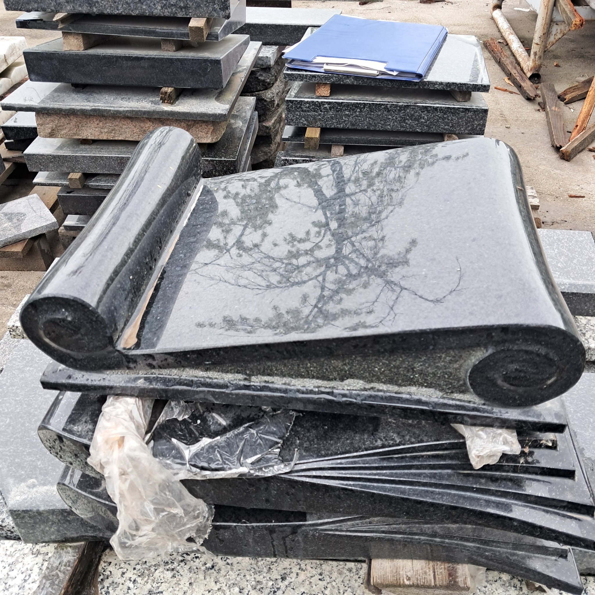 Stos czarnych, polerowanych płyt kamiennych z zawiniętymi krawędziami, na jednej z nich widoczne odbicie drzewa, całość częściowo zabezpieczona folią.